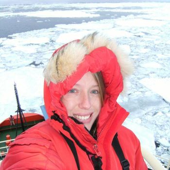 Woman in red parka smiles aboard ship in icy waters.