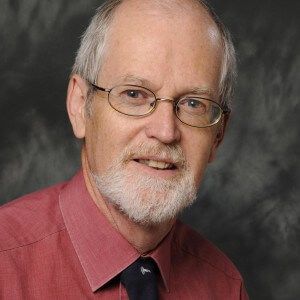Man with glasses and white beard wearing a red shirt and dark tie, smiling in front of a neutral backdrop.