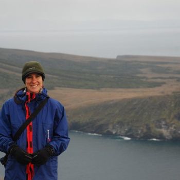 Person in blue jacket and beanie smiles, standing on a cliff overlooking the ocean and hills under a cloudy sky.
