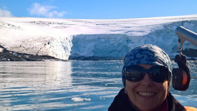 Woman smiling, wearing sunglasses and hat, in front of a glacier and icy water.