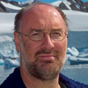 Man with glasses and beard, in Antarctica, against snowy mountains and icebergs.