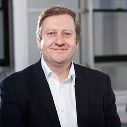 Man in a dark suit and white shirt smiles, indoors.