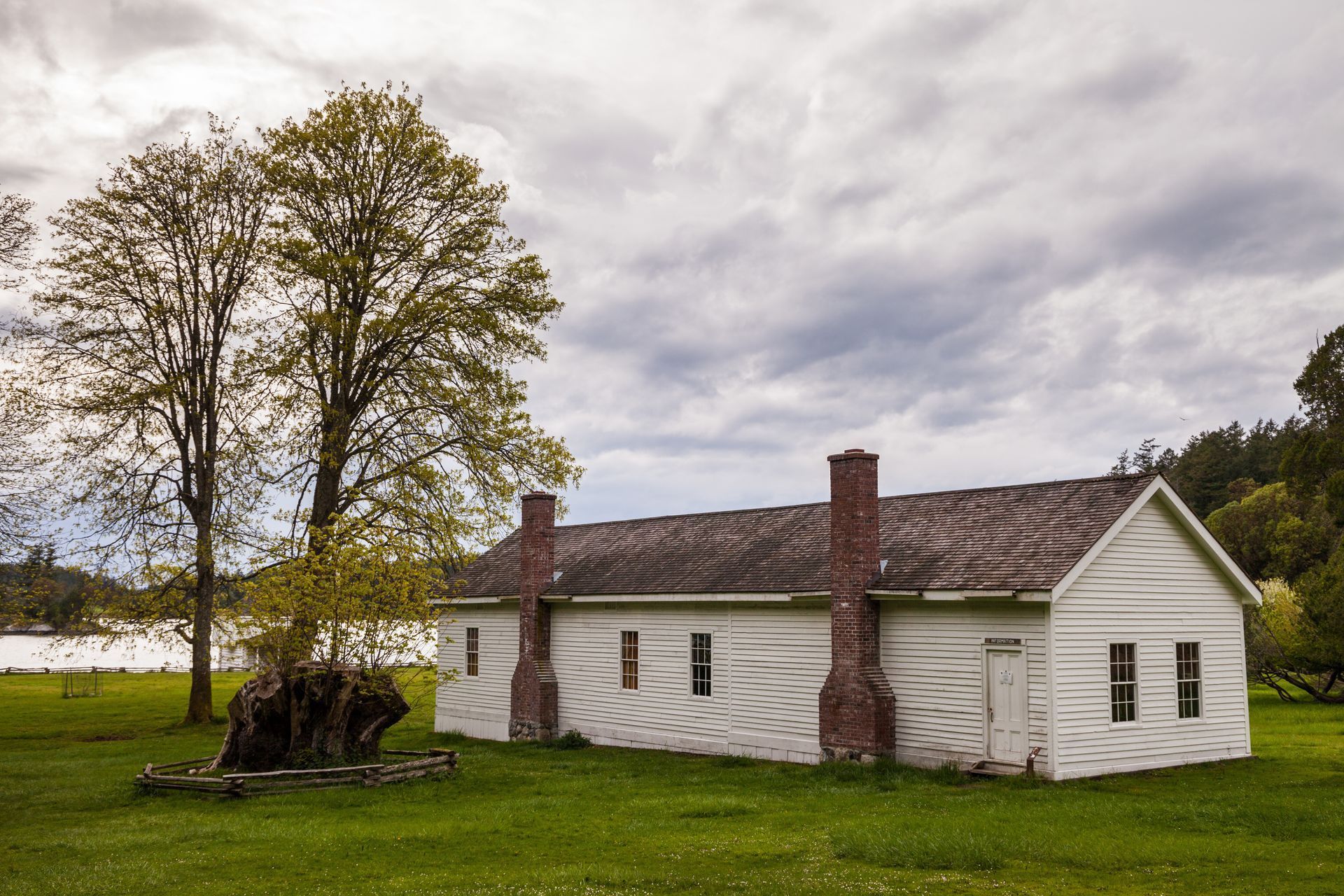 Small white farmhouse with two chimneys beside a leafless tree in a grassy field under a cloudy sky