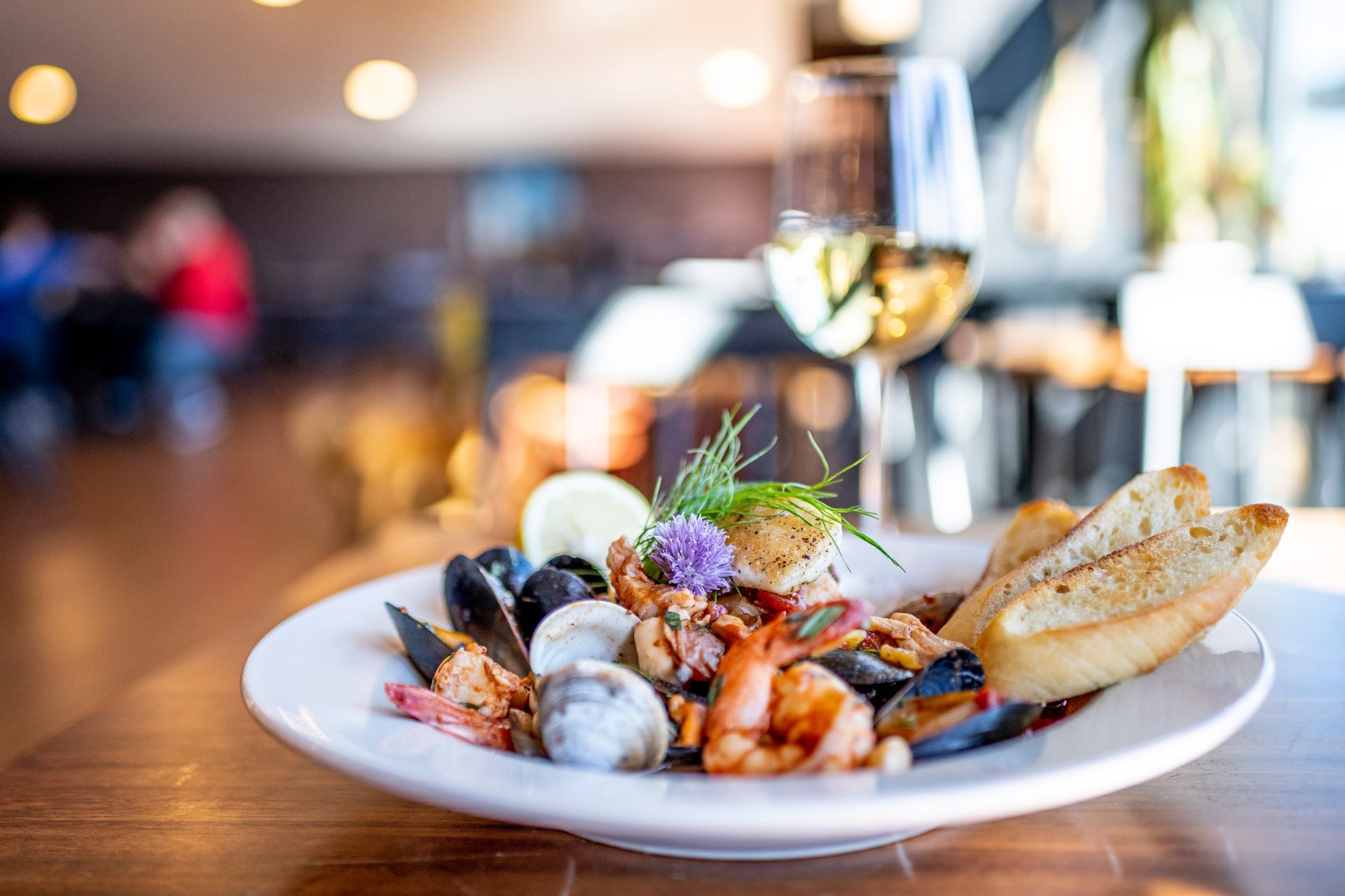 Seafood pasta with mussels, shrimp, and bread on a white plate beside a glass of white wine