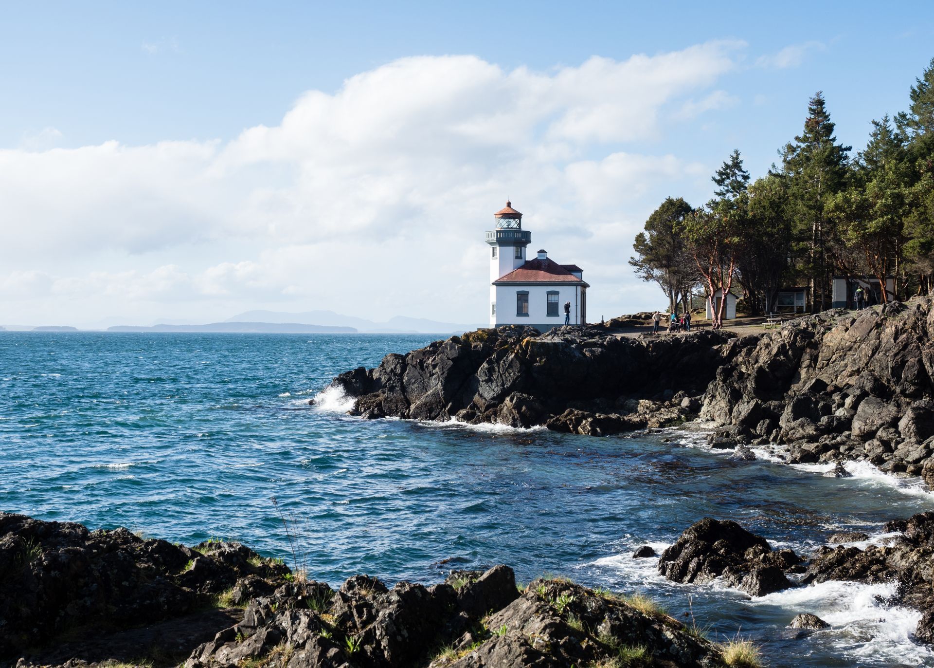 Lighthouse on a rocky coastline with blue ocean waves and pine trees under a clear sky