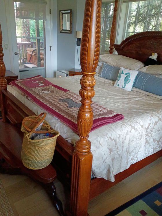 Wooden four-poster bed with white bedding, red runner, and woven basket in a bright bedroom