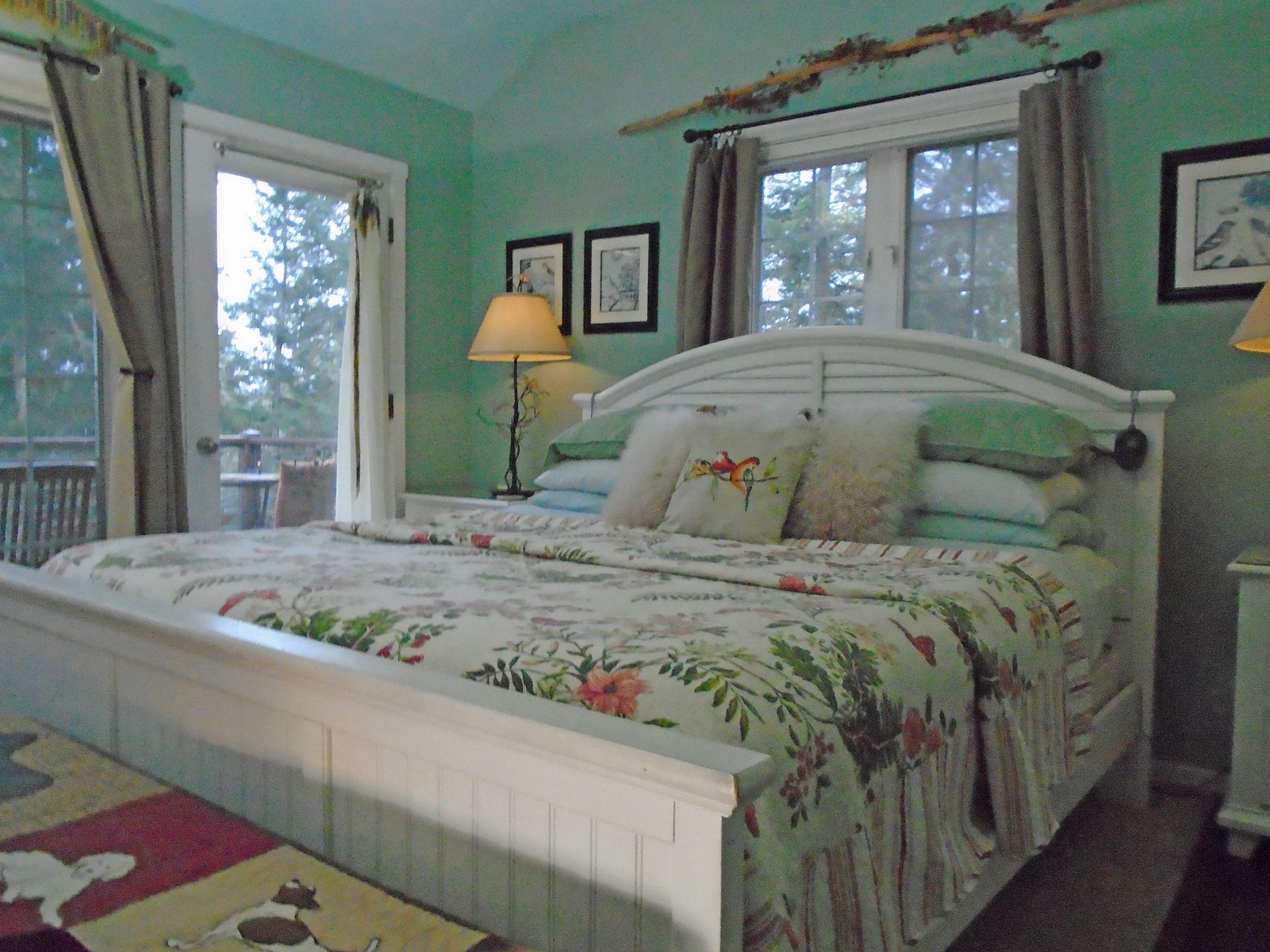 Cozy bedroom with a white bed, floral quilt, green walls, and windows with curtains and a lamp.