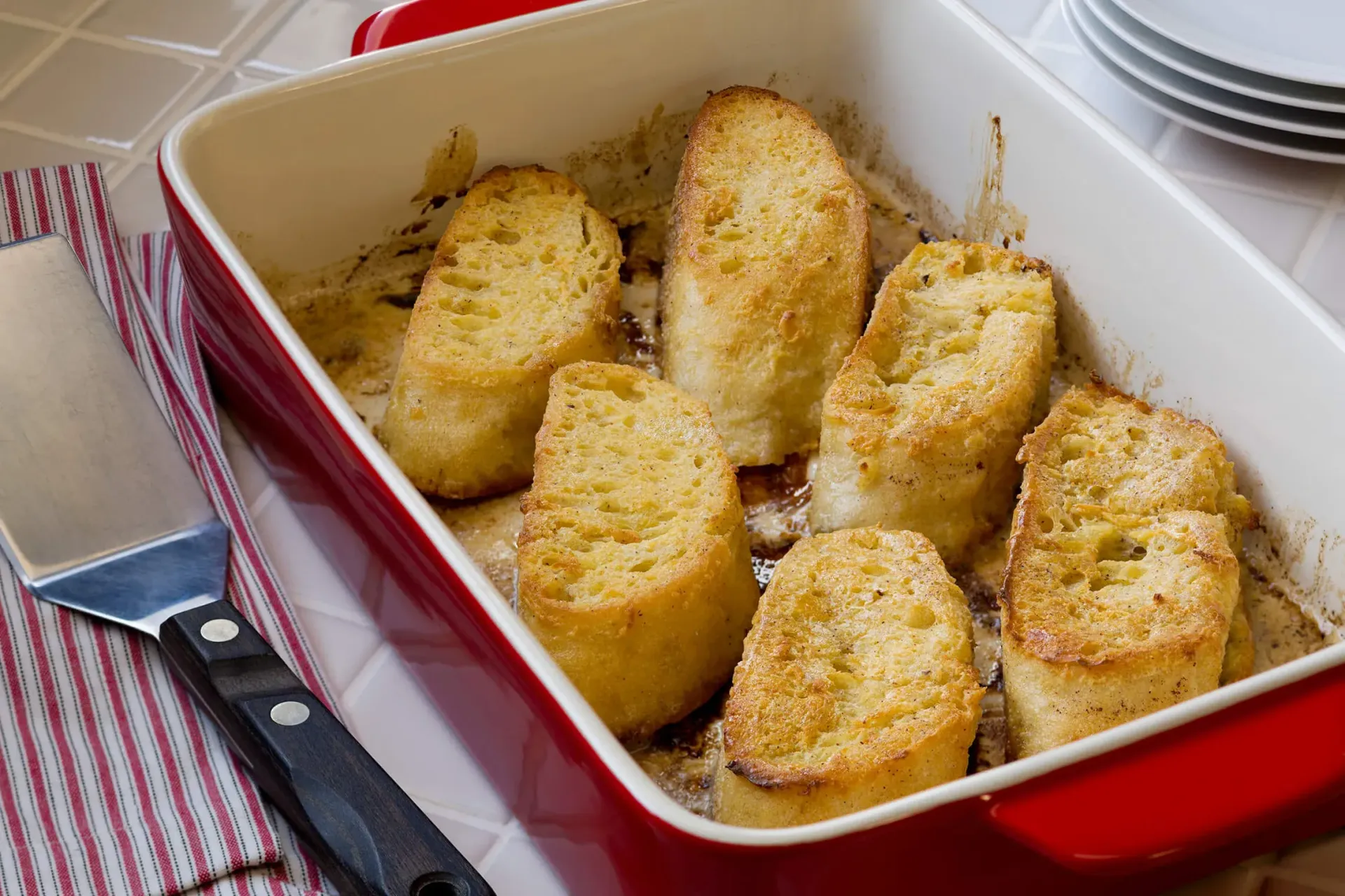 Red baking dish with six golden bread slices on a checkered cloth, with a fork beside it