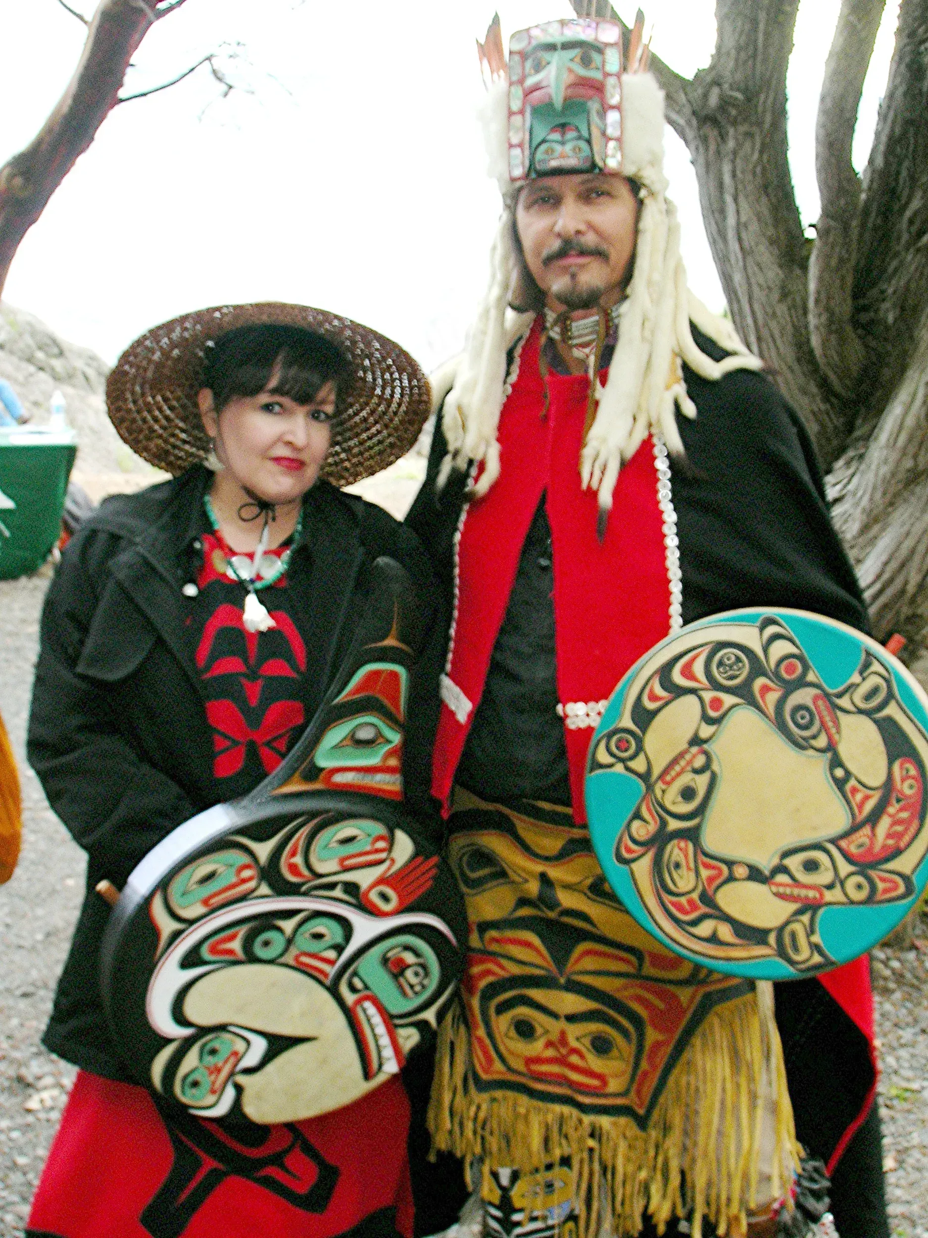 Two people in traditional Indigenous regalia holding painted shields and wearing ceremonial headdresses outdoors