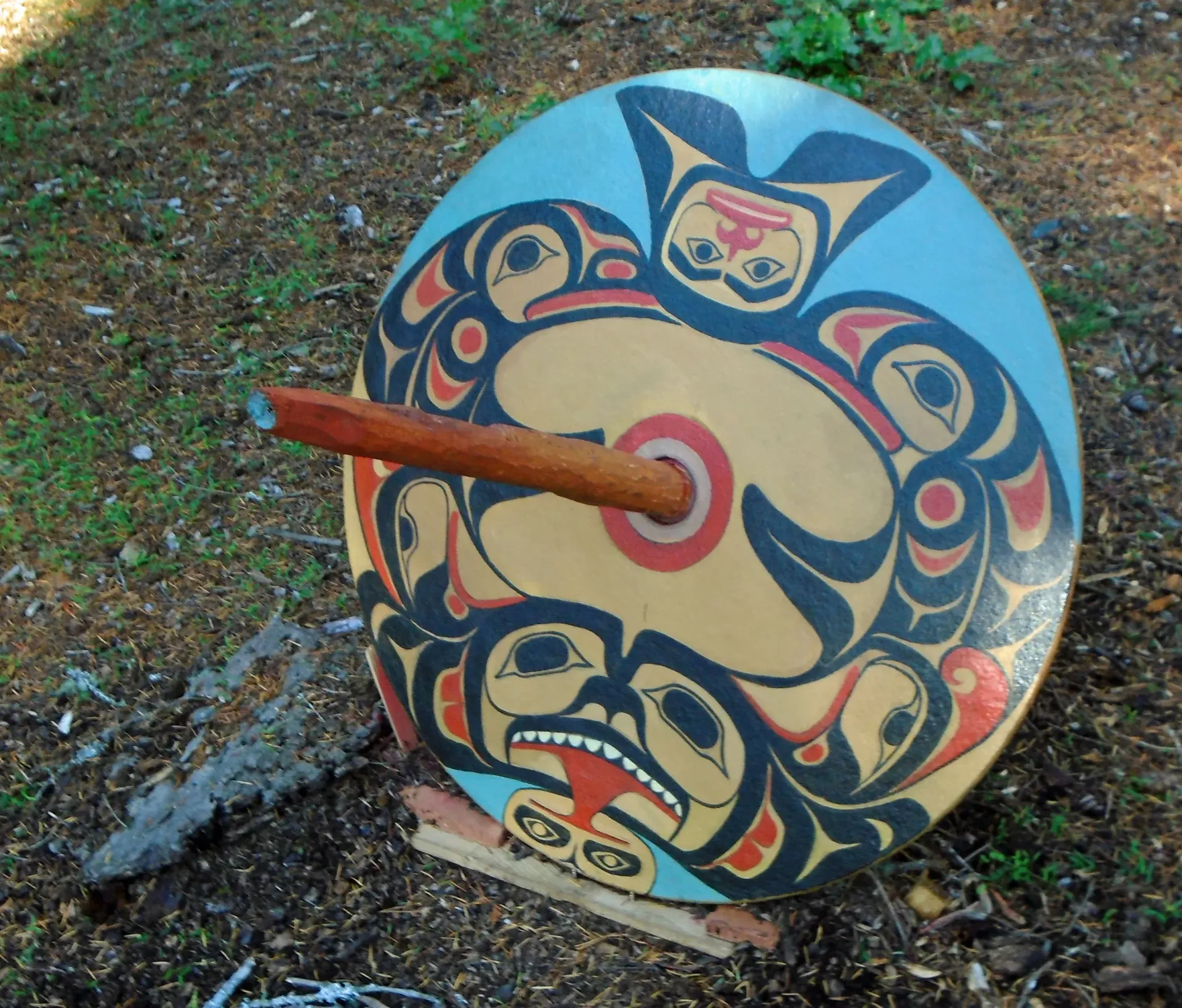 Decorated round drum with a wooden beater sticking through the center, set on the ground outdoors.