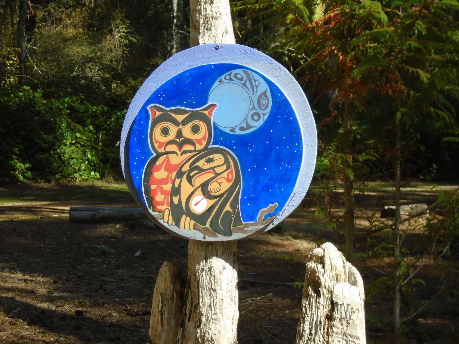 Hand-painted owl on a blue circular sign mounted on wooden posts in a wooded area