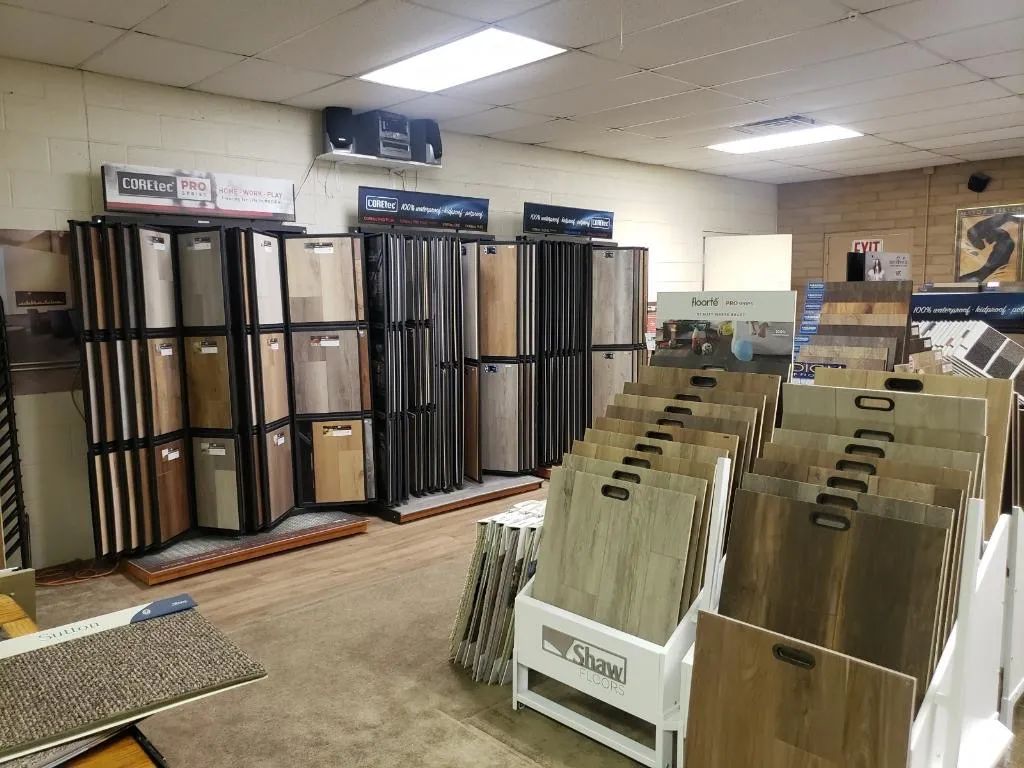 Store interior with stacked floor mats and racks of sample flooring displays under fluorescent lights