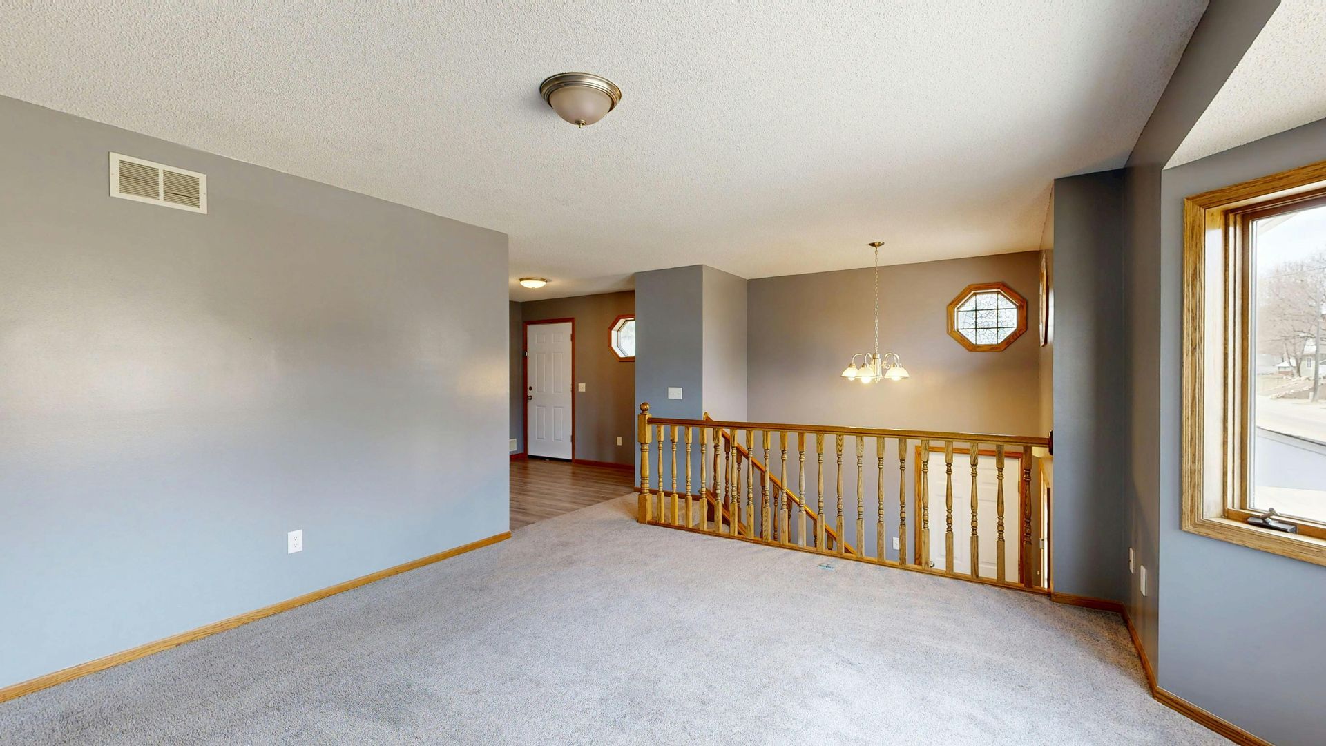 Empty beige living room with carpet, stair railing, and a doorway to the right.