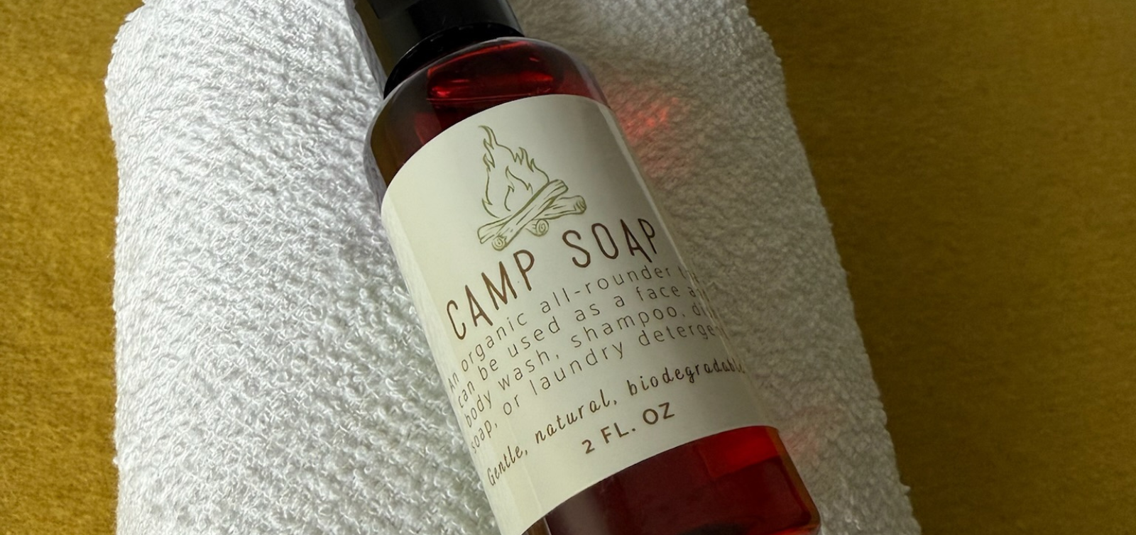 Camp Soap bottle on a white towel against a yellow background. The bottle has a black cap.