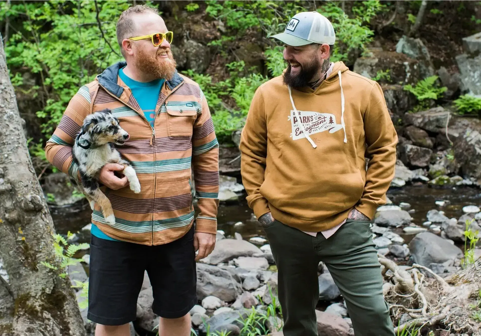 Two men wearing outdoor inspired apparel looking at each other. One is holding a dog