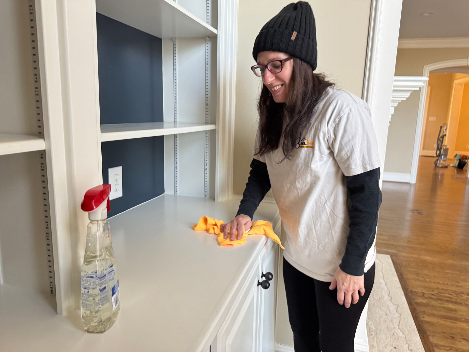 Woman cleaning a white shelf with a spray bottle and cloth; indoor setting.