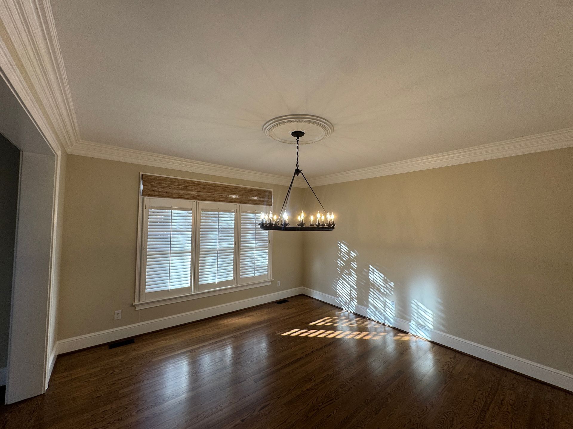 Empty dining room with chandelier, window, and hardwood floors. Sunlight casts shadows on the wall.