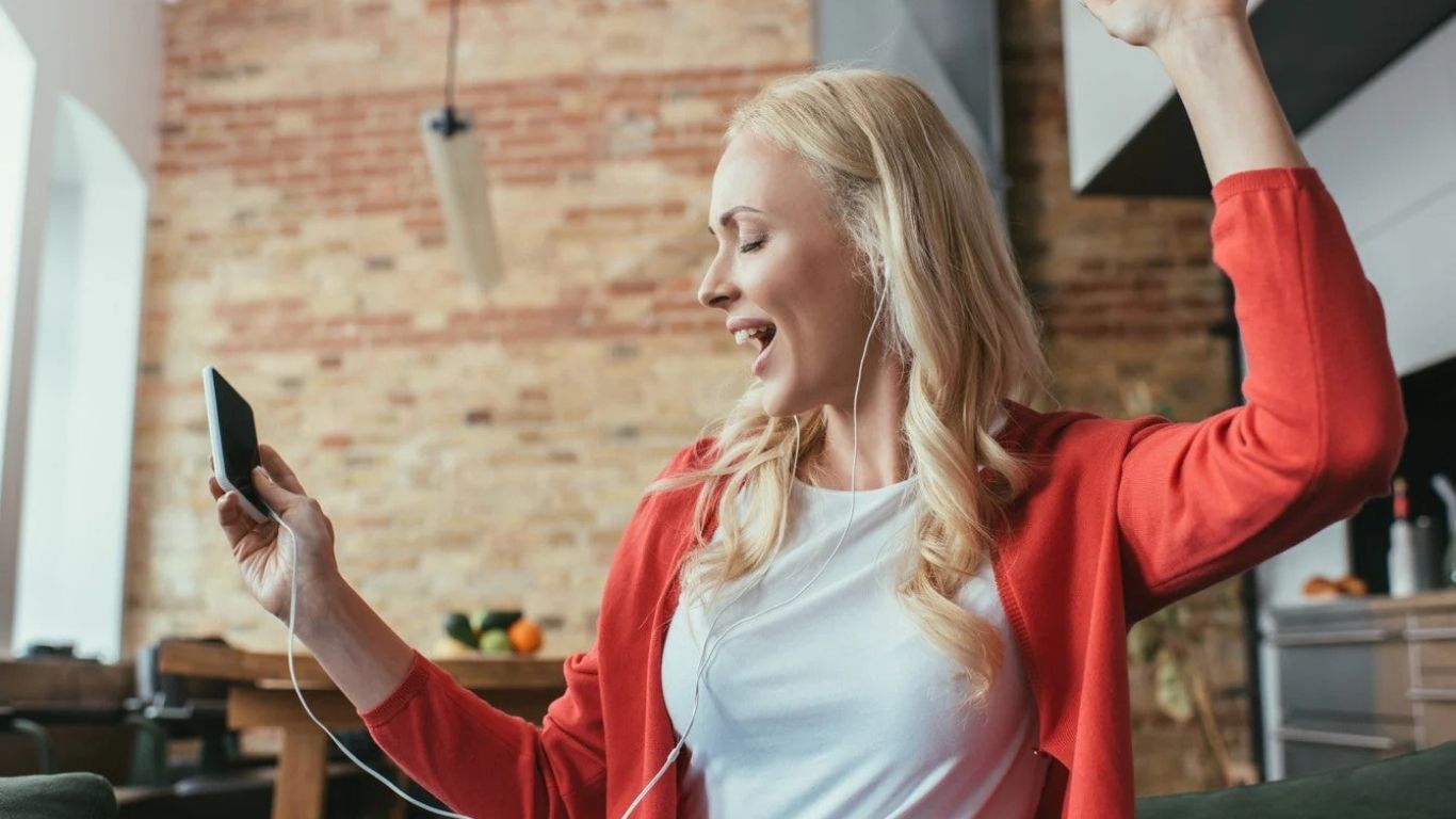 Woman with blonde hair, wearing a red cardigan, listening to music on her phone, and celebrating with her arm raised in a room with exposed brick.