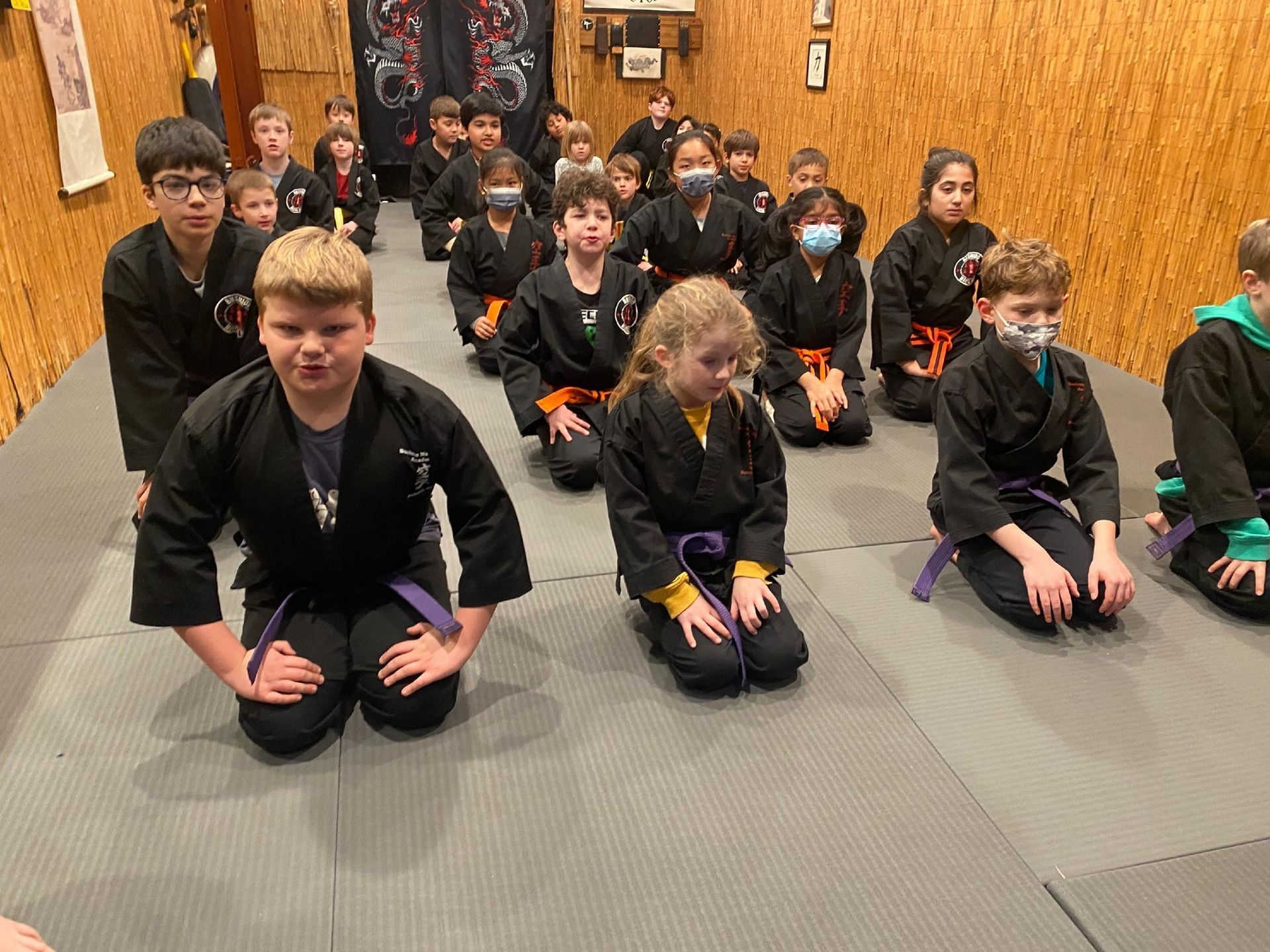 A group of children in karate uniforms are kneeling on the floor in a gym.