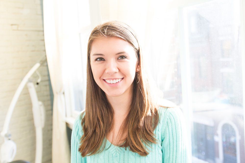 A woman in a blue sweater is smiling in front of a window.