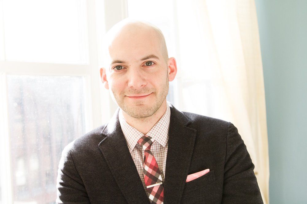 A bald man in a suit and tie is smiling in front of a window.