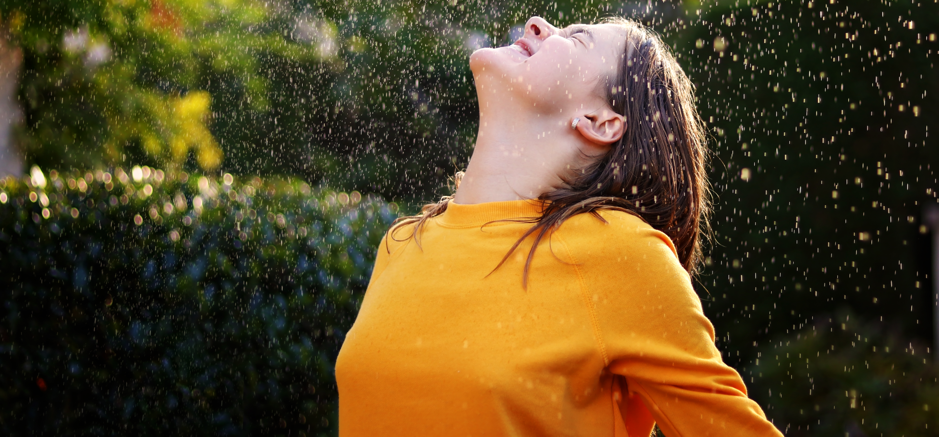 A woman in a yellow sweater is standing in the rain with her arms outstretched.