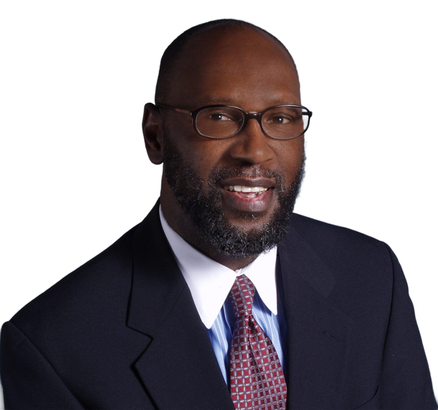 Man in a dark suit and patterned tie, wearing glasses, smiling against a white background