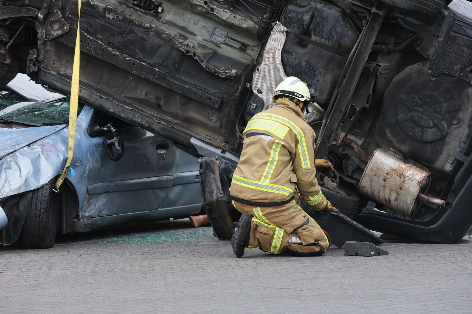 Firefighter kneels beside a crashed car pinned under an overturned truck on a road.