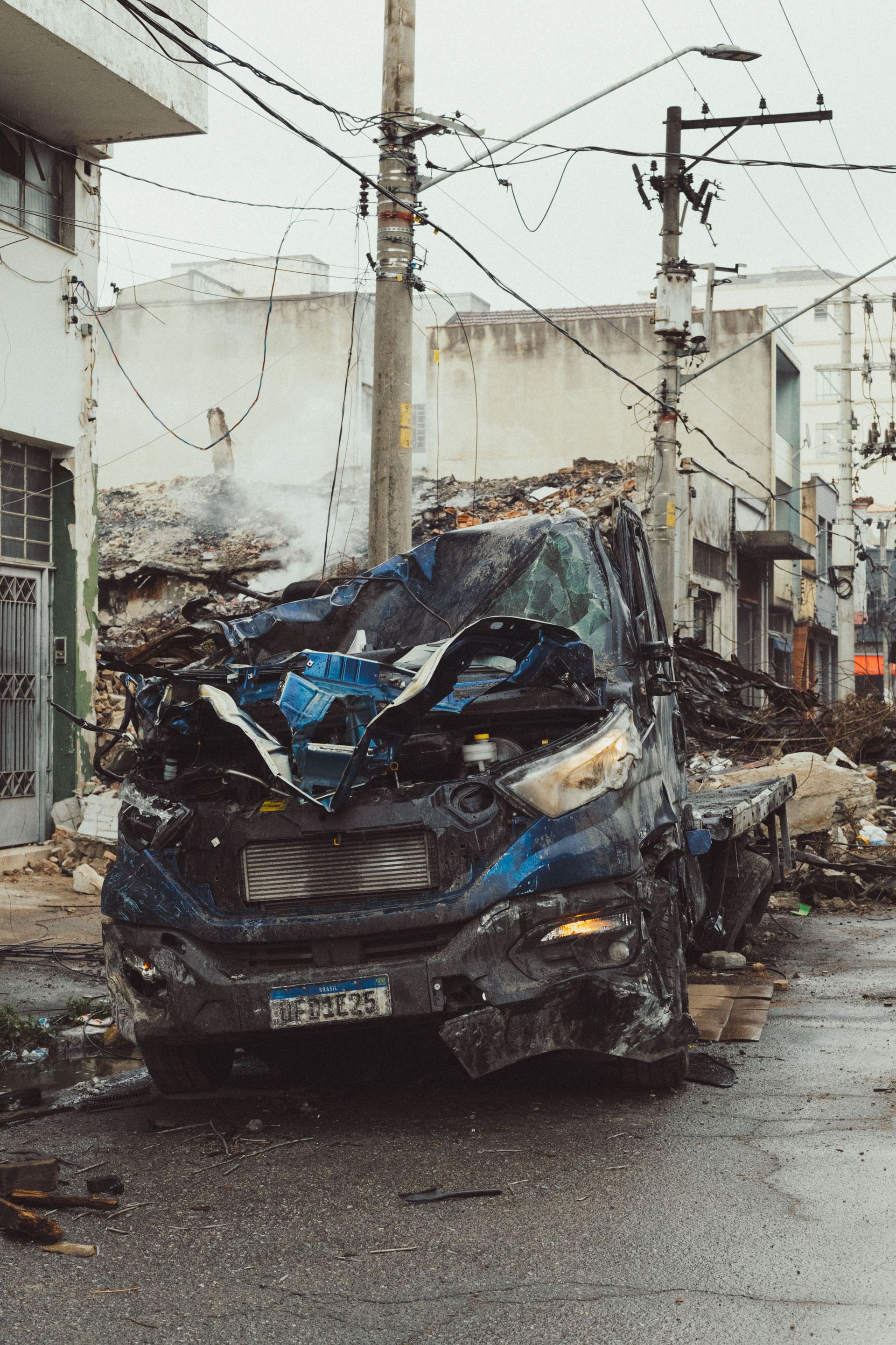 Severely damaged blue car crumpled on a wet street in a residential area, with power lines overhead