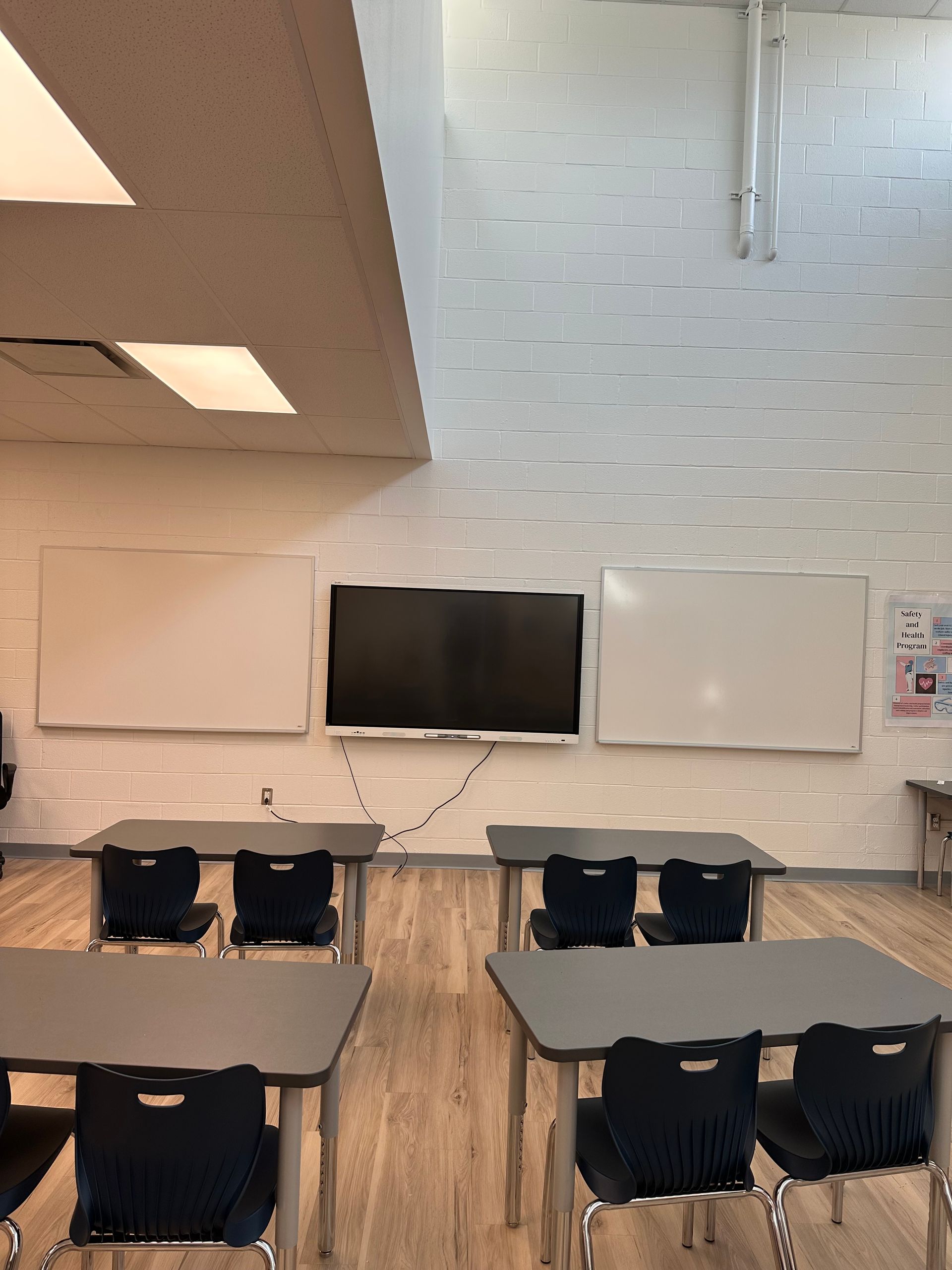 Empty classroom with desks, chairs, a wall-mounted TV, and whiteboards on a white brick wall.
