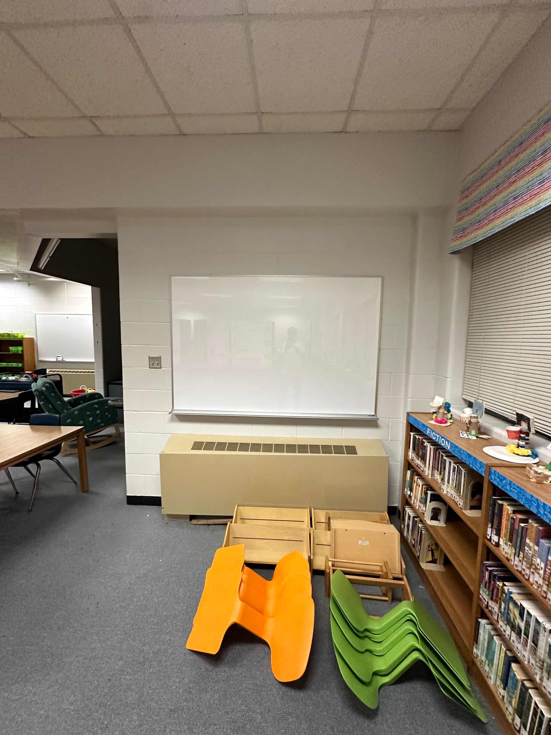 Classroom corner with stacked chairs, desks, bookshelves, and boxes under a whiteboard.