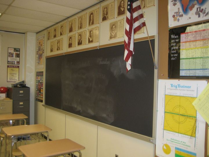 Empty classroom with a large chalkboard, desks, wall posters, and a hanging American flag.