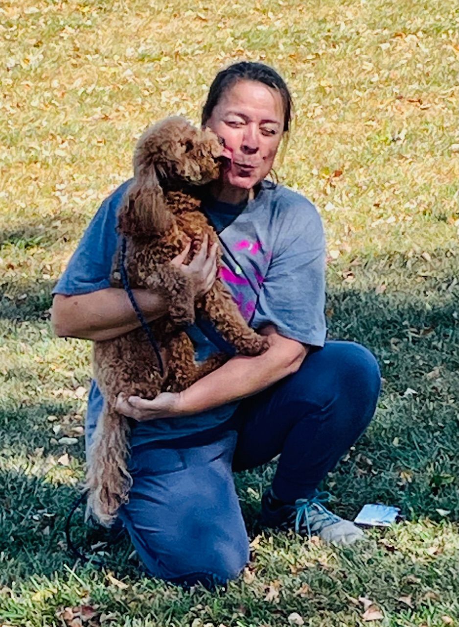 Woman kneeling, hugging and kissing a brown poodle in a grassy area.