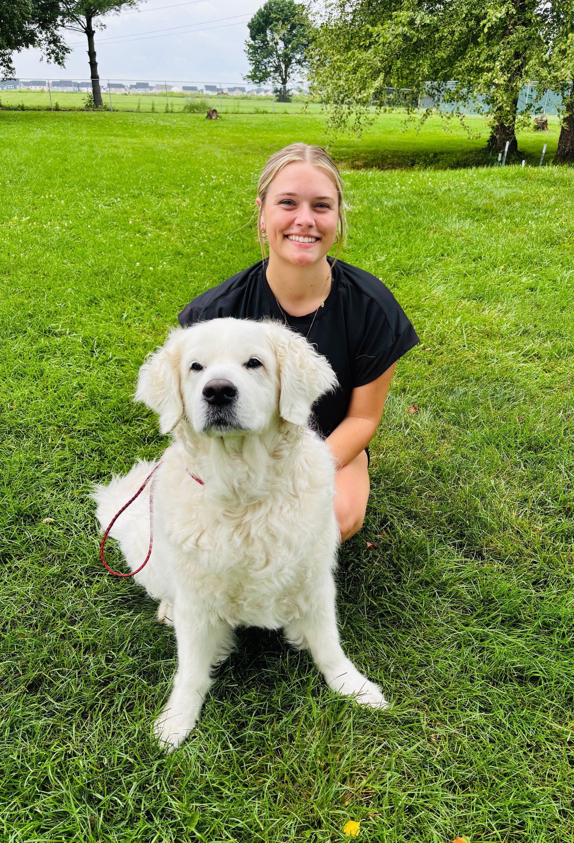Woman kneeling next to a white fluffy dog on a grassy field, both looking at the camera.