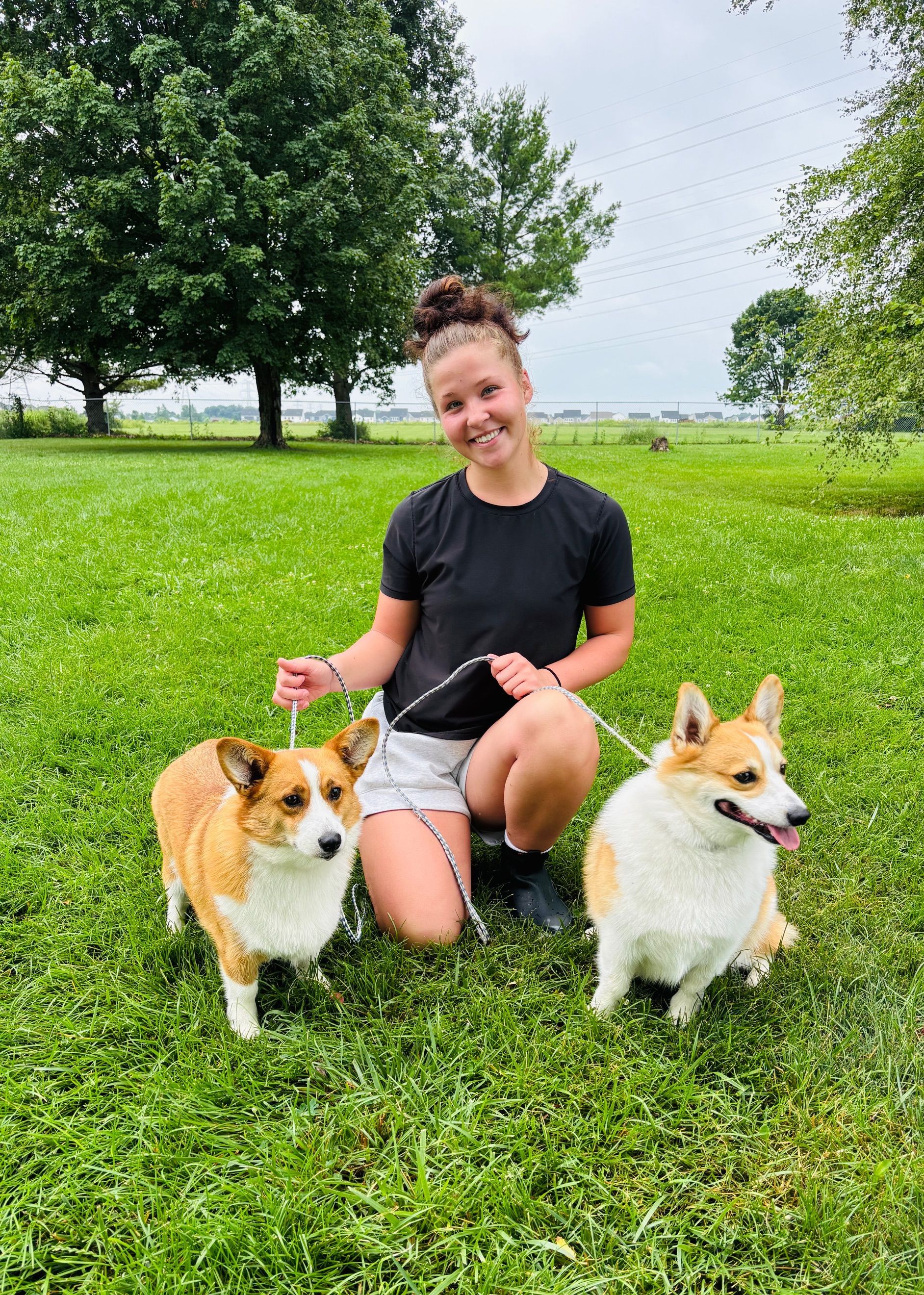 Woman kneels with two corgi dogs on a grassy lawn. The dogs are tan and white. She is smiling.