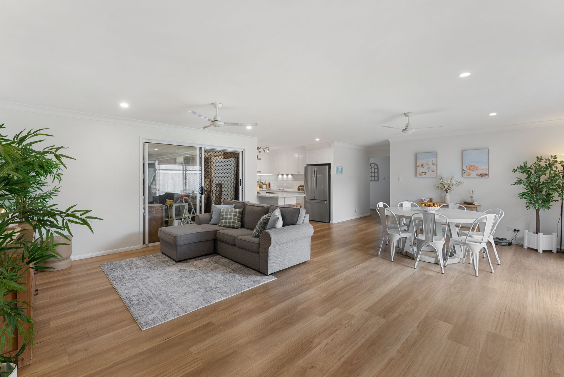 An open-plan living and dining room with a grey sofa, area rug, and white dining table set on light wooden floors.