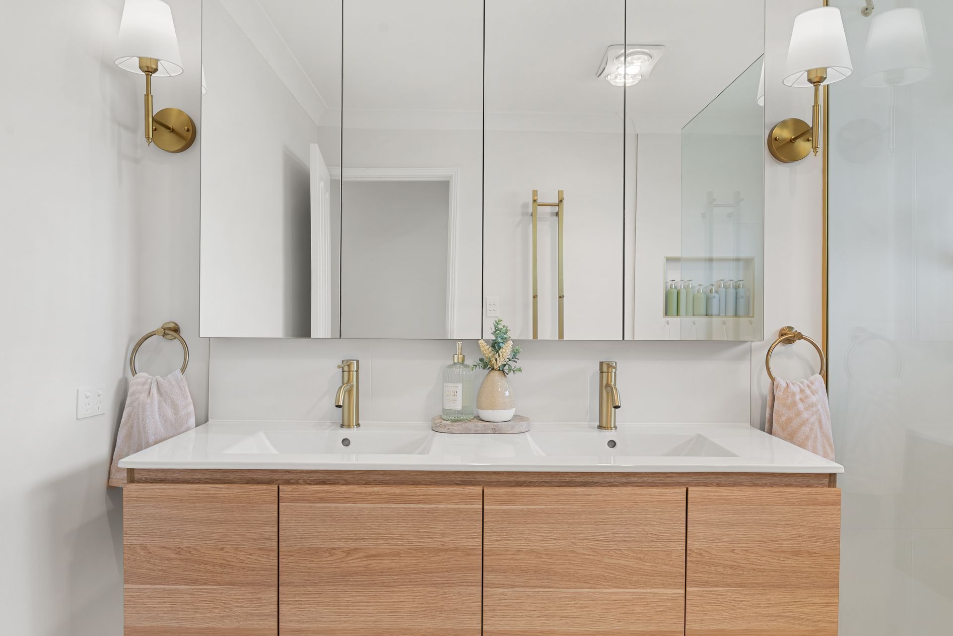 Modern white bathroom with double vanity, brass faucets, large mirror, and wood cabinet.