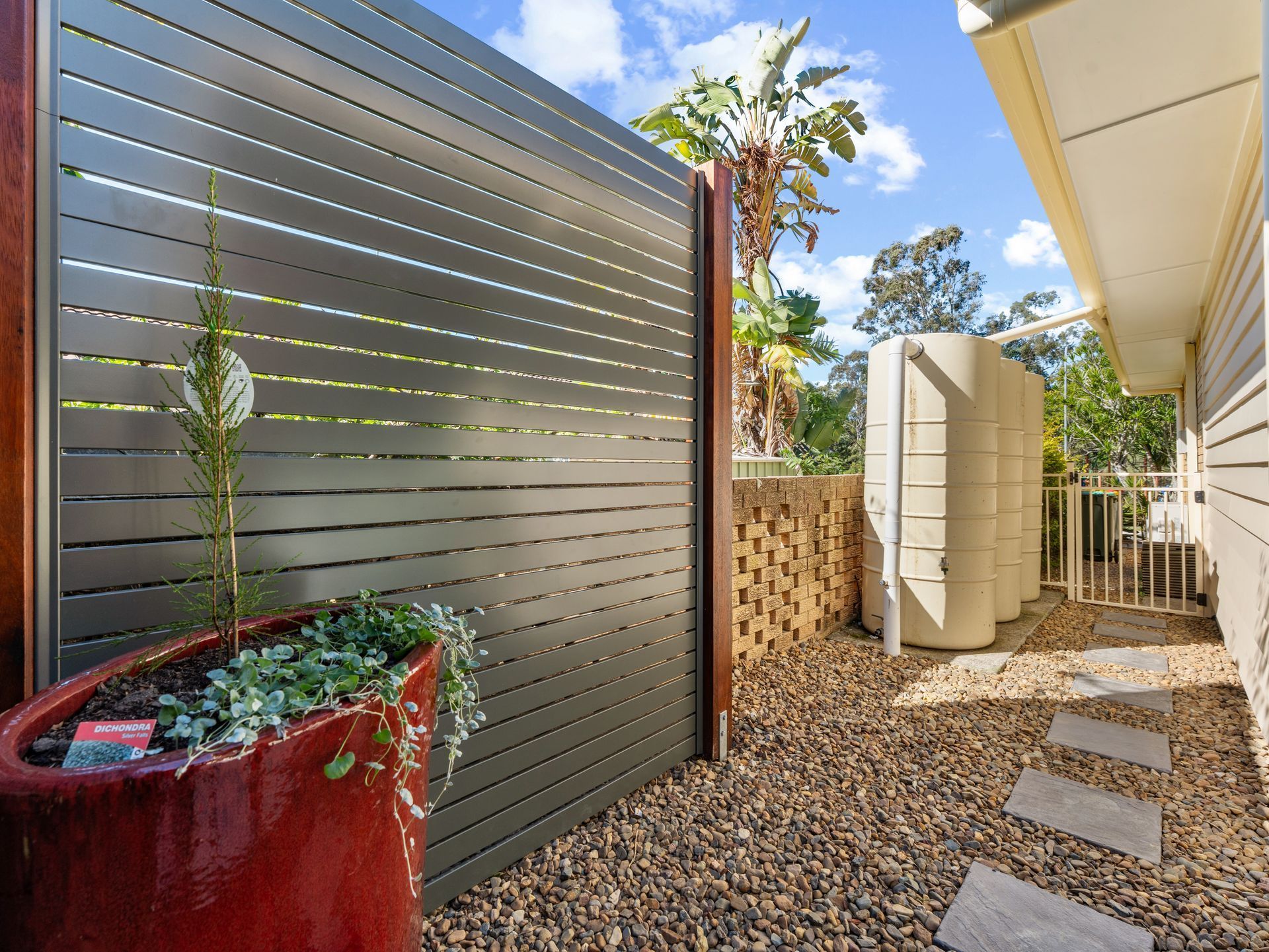Modern home patio with gray privacy fence, red planter, stepping stones, and gravel pathway under bright sky