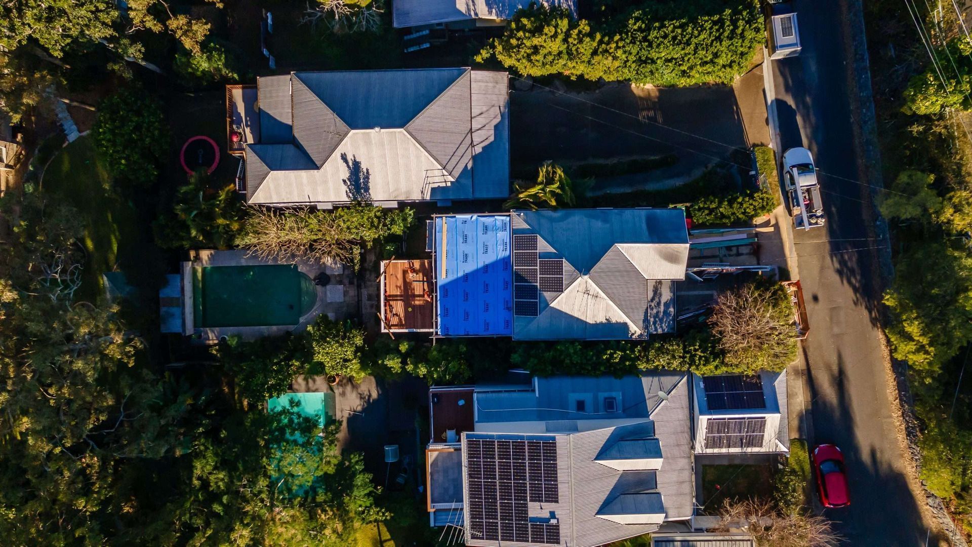 Aerial view of several houses with blue roofs along a tree-lined street and parked cars