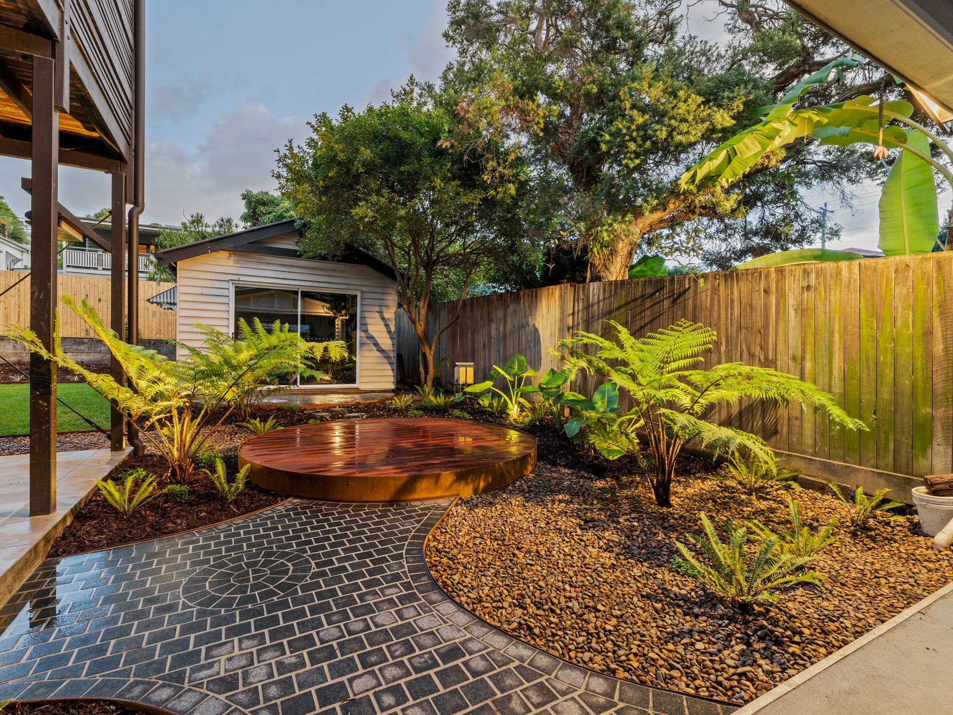 Backyard garden with cobblestone path, ferns, a circular wooden patio, and a shed.