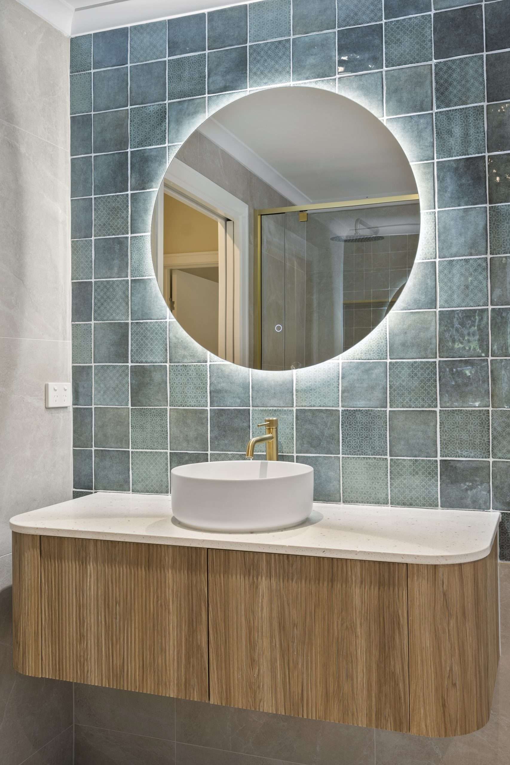 Bathroom with round backlit mirror, wood vanity, vessel sink, and blue square tiles.