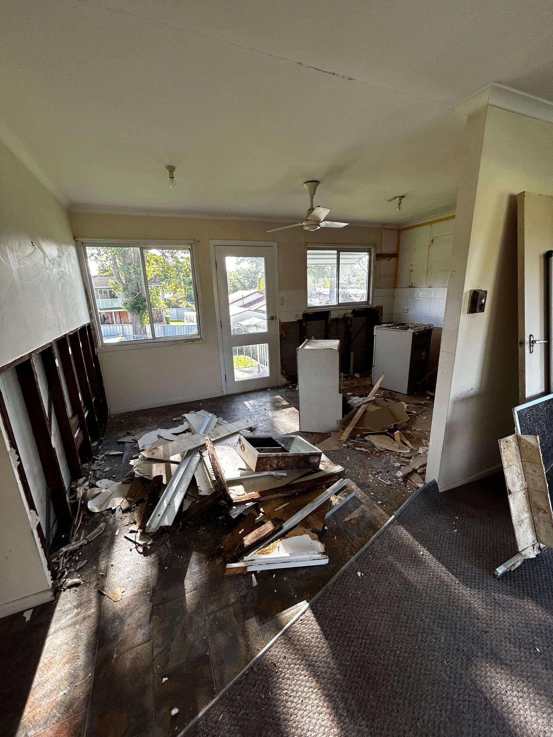 Interior view of a room undergoing demolition, debris scattered across the floor. Windows, doorway, and ceiling fan are visible.