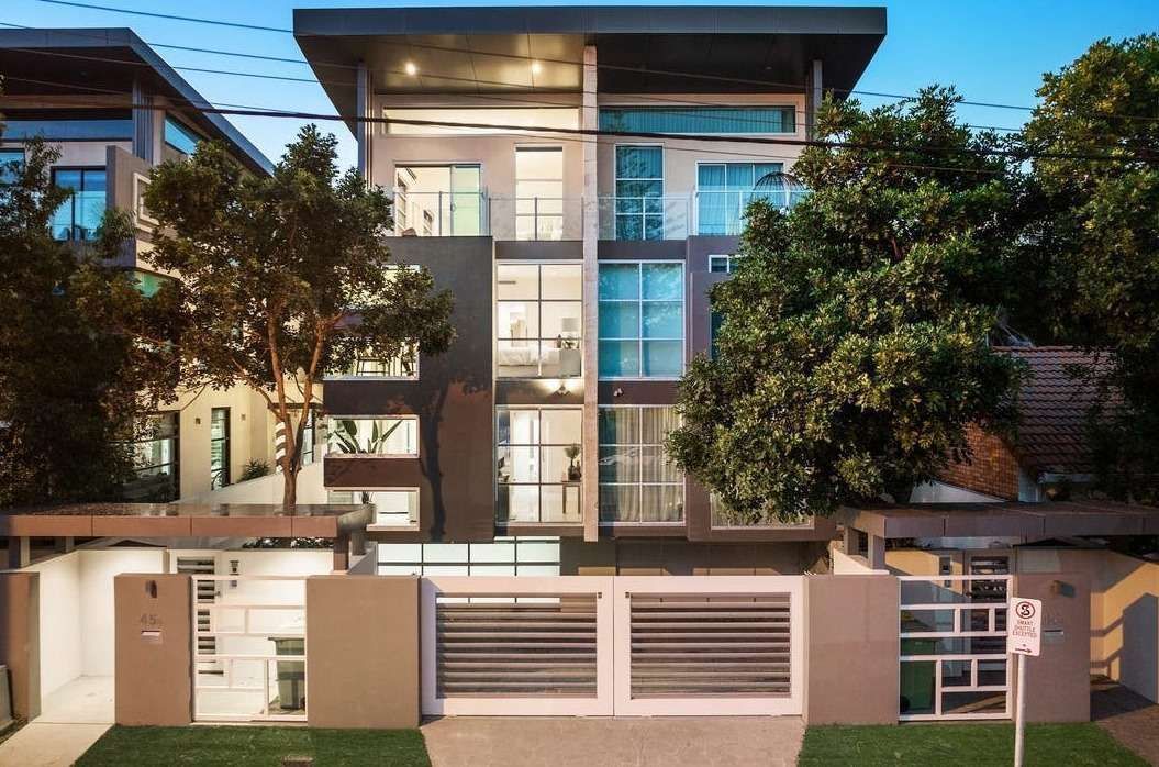 Modern two-story house with glass balconies, white walls, and a front gate surrounded by trees