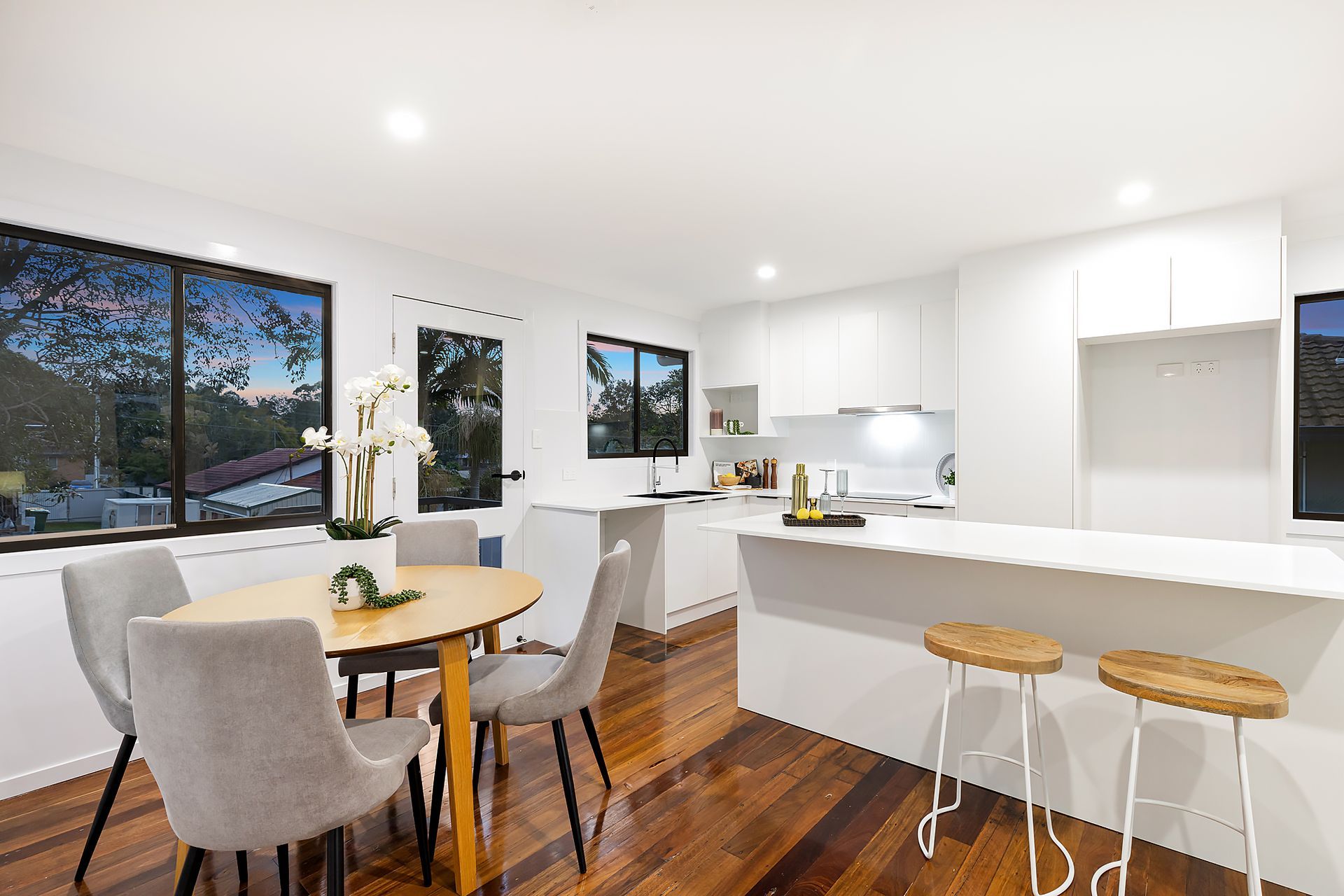 Bright, modern kitchen and dining area with wooden floors. Features a table with chairs, a breakfast bar, and a window view.