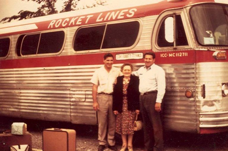 Three people posing in front of a vintage Rocket Lines bus with luggage nearby