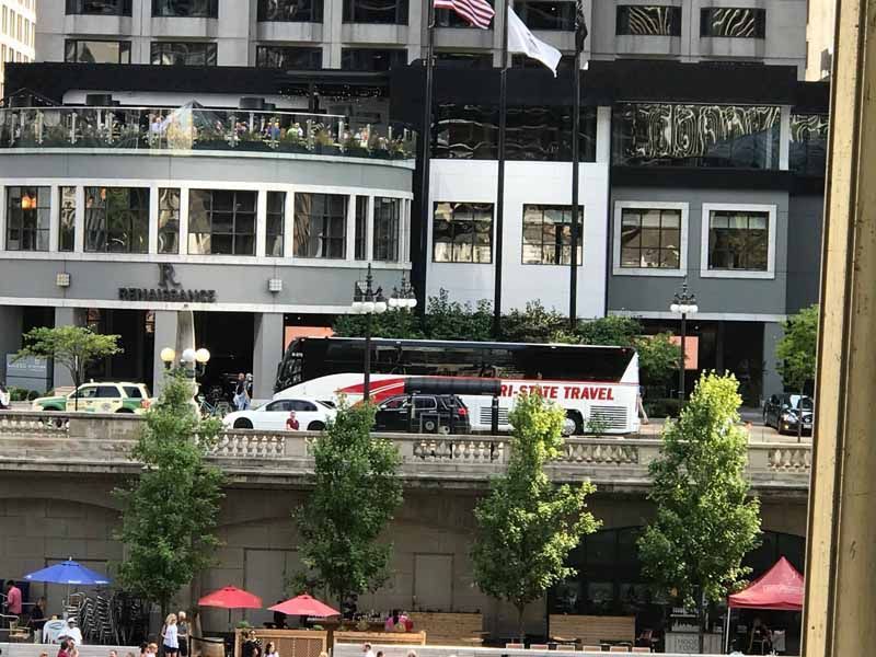 City waterfront scene with a white tour bus and people walking beside a concrete embankment.