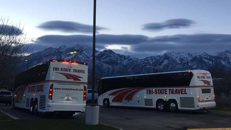 Two white TRI-STATE TRAVEL buses parked at dusk with snowy mountains in the background