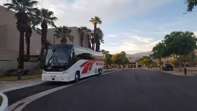 White tour bus parked on a palm-lined street at sunset near a large building