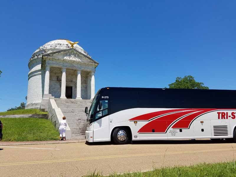 White memorial with columns on a grassy hill beside a parked white coach bus under a blue sky
