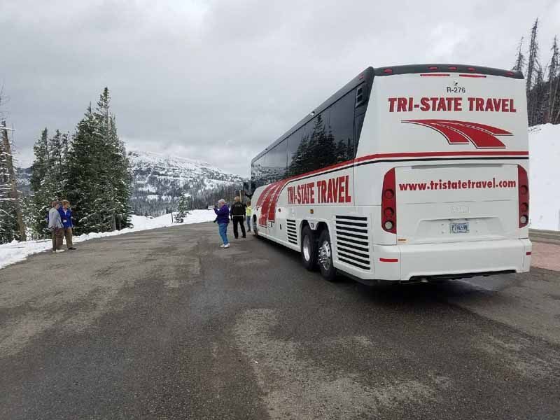 White Tri-State Travel coach bus on a snowy mountain road with people and trees nearby